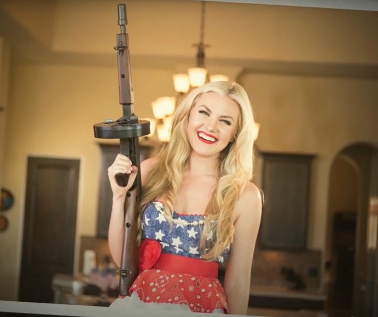 Image still from an AMA promotion video. Erika is standing in a southwestern-style home wearing a star-spangled outfit. Her hair is long, flowing, and blond. Her lips are blood red. She is smiling and holding a "Tommy" gun.